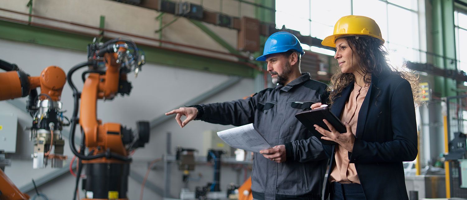 Two people wearing protective helmets walk through a workshop and look at the work processes